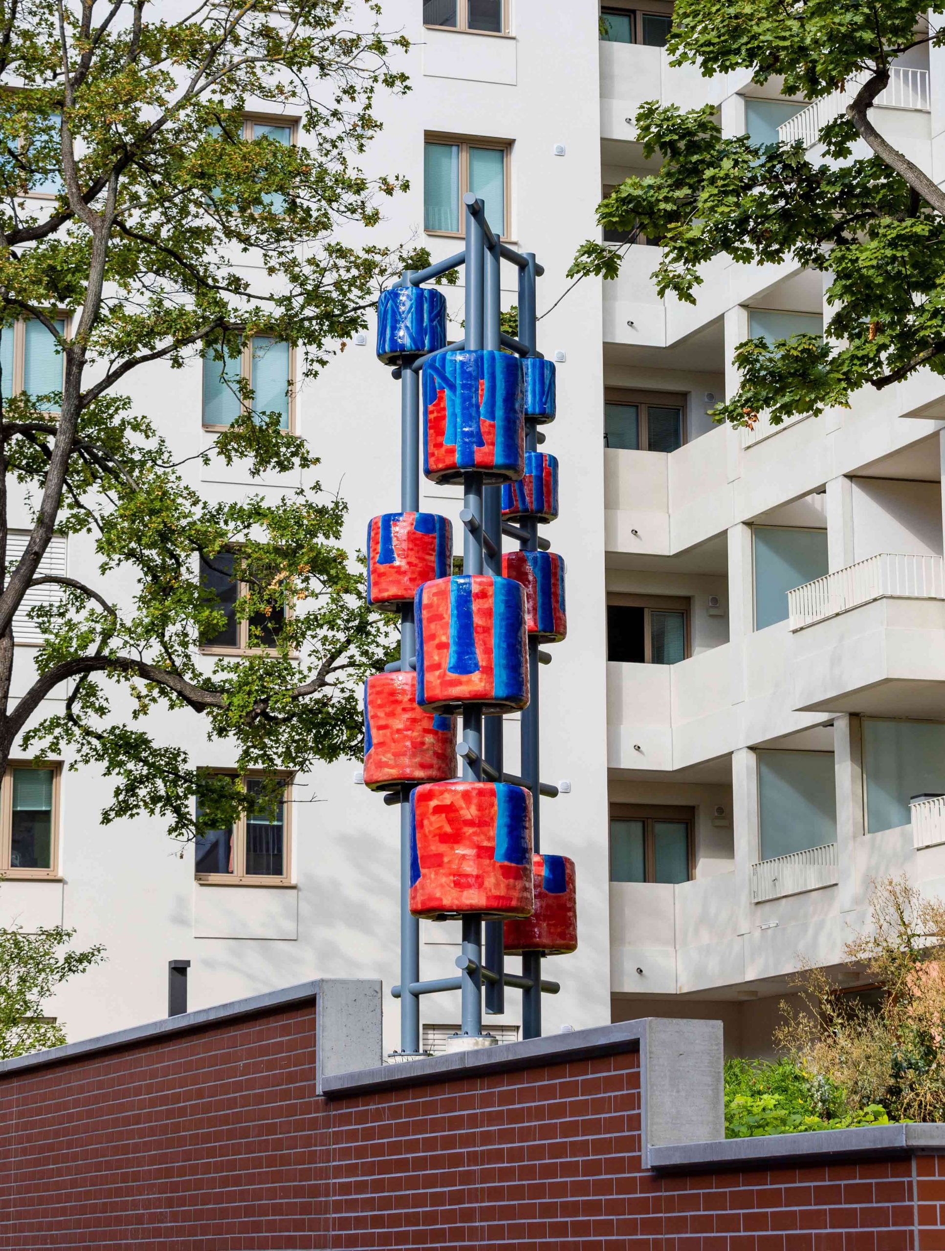 a large red and blue ceramic sculpture with steel supports and apartment buildings and trees in the background 