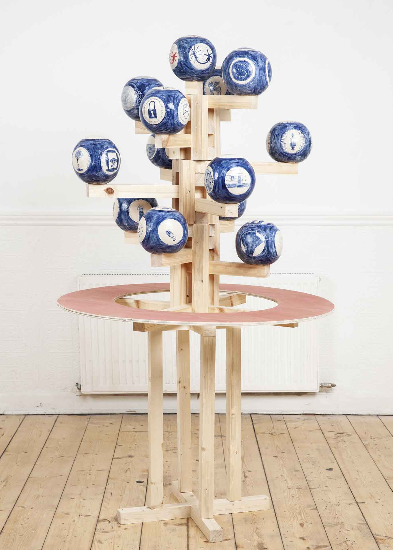Blue and white ceramic spheres assembled on a wooden frame not unlike a christmass tree, inside a white gallery space with unvarnished wooden floors. 