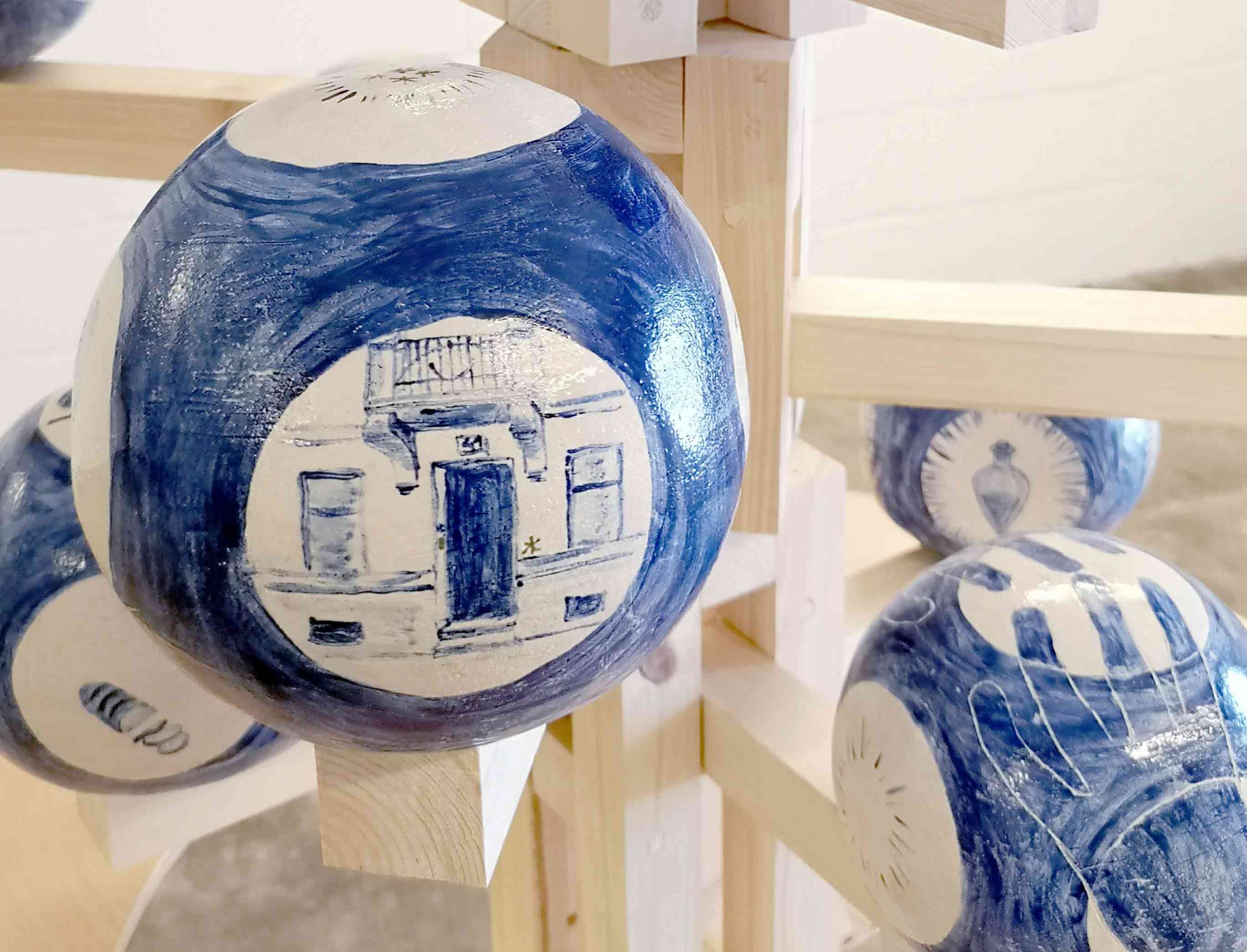 A close up photograph of one of the cermaic spheres in the previous photo, showing the painted surface which has an image of a doorway underneath a iron balcony, painted in blue on a white background. the other spheres are visible at the edge of the frame. 